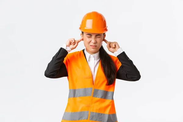 Asiática gerente de empresa do sexo feminino sentindo desconforto, engenheiro chefe fechou as orelhas e sorrindo de ruído irritante, inspecionar o trabalho na zona de construção, som horrível de reparação, usar capacete de segurança — Fotografia de Stock