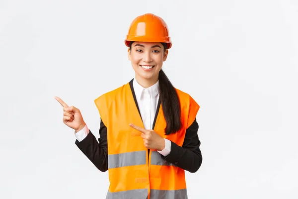 Construcción, construcción y concepto industrial. Sonriente asiática arquitecta en casco de seguridad, ropa reflectante apuntando dedo esquina superior izquierda, mostrando proyecto en el lugar de trabajo, fondo blanco — Foto de Stock