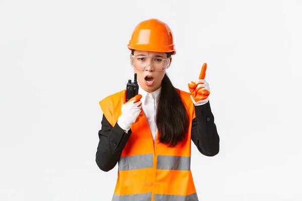 Serious-looking asiático engenheiro industrial feminino, técnico em capacete de segurança e equipe de construção de comando uniforme com walkie-talkie, explicar ou repreender alguém com telefone de rádio — Fotografia de Stock