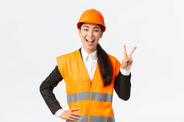 Gelukkige vrolijke Aziatische vrouwelijke Aziatische architect in veiligheidshelm en reflecterende jas, toont vrede teken en glimlach, verzekerd in de overwinning van haar bedrijf, winnende tender op bouwwerkzaamheden — Stockfoto