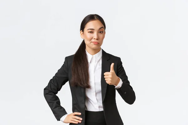 Ευχάριστη γυναίκα επιχειρηματίας, κυρία αφεντικό με μαύρο κοστούμι ικανοποιημένη με τη δουλειά σας, δείχνει τους αντίχειρες προς τα πάνω και νεύμα με έγκριση, έπαινος μεγάλη δουλειά, καλά κάνει, στέκεται λευκό φόντο — Φωτογραφία Αρχείου