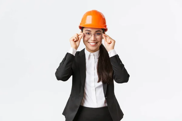 Χαμογελώντας επιτυχημένη επιχειρηματίας σε μαύρο κοστούμι και κράνος ασφαλείας επισκεφθείτε τον κατασκευαστικό τομέα για την παρακολούθηση της εργασίας, χαμογελώντας ικανοποιημένοι, να θέσει σε γυαλιά, μηχανικός κτίριο διαχείριση των εργαζομένων, λευκό φόντο — Φωτογραφία Αρχείου