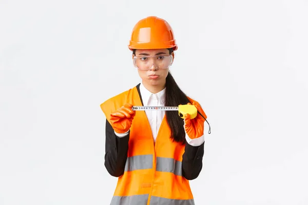 Teleurgesteld sombere vrouwelijke Aziatische bouwkundig ingenieur, architect toont kleine maat op tape maatregel, pruilen boos met metingen, staan ontevreden in veiligheidshelm, witte achtergrond — Stockfoto