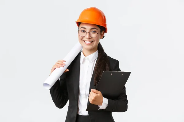 Sucesso asiático arquiteto feminino em terno de negócios e capacete segurando plantas e prancheta com notas, inspetor olhando para obras de construção, sorrindo para a câmera, fundo branco — Fotografia de Stock