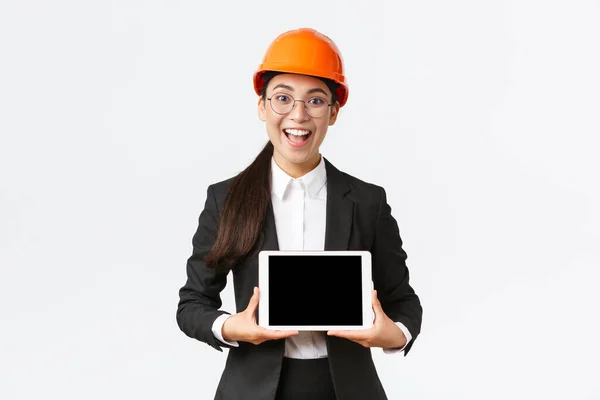 Aufgeregt und erstaunt lächelnd asiatische Bauingenieurin, Architektin in Schutzhelm und Businessanzug, zeigt digitalen Tablet-Bildschirm mit erstauntem Gesichtsausdruck, stehender weißer Hintergrund — Stockfoto