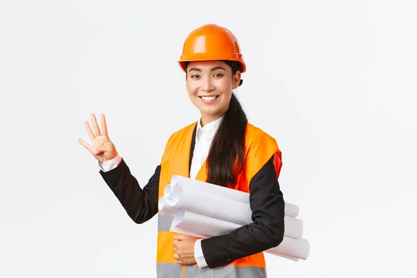 Glimlachend zelfverzekerd aziatische vrouwelijke ingenieur, bouwmanager in veiligheidshelm, dragen blauwdrukken, met vier vingers, zorgen voor het gebouw klaar in de tijd, staande witte achtergrond upbeat — Stockfoto