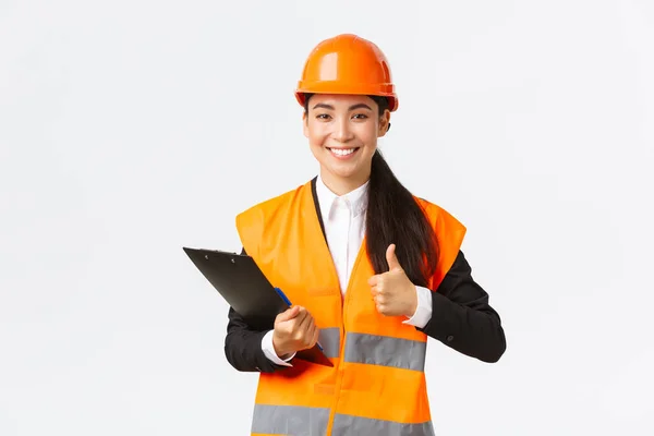 Zadowolona azjatycka inżynier budowlana zadowolona z wyników inspekcji, noszenia odzieży ochronnej i kasku na placu budowy, zapisywania notatek i pokazywania kciuków w aprobacie — Zdjęcie stockowe