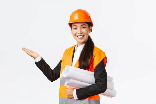 专业的亚洲女建筑师微笑，安全帽工程师介绍建筑工程，用左手指著携带设计图，讲演，介绍建筑图表或平面图 — 图库照片