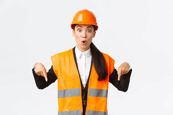 Incroyable et intéressé asiatique femme architecte, femme industrielle en veste réfléchissante, casque de sécurité pointant les doigts vers le bas et l'air impressionné, parler de construction de bâtiments, fond blanc — Photo