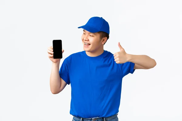 Tecnologia, compras on-line e conceito de transporte. Sorrindo satisfeito asiático entregador homem em azul uniforme, mostrando polegares para cima e olhando satisfeito na tela do telefone móvel, recomendo app, fundo branco — Fotografia de Stock