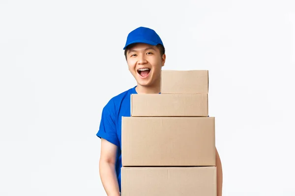 Online-Shopping, schnelles Versandkonzept. Optimistisch lächelnder asiatischer Zusteller in blauer Kurieruniform hält Kisten mit Bestellungen, trägt Pakete zum Haus des Kunden, steht vor weißem Hintergrund fröhlich — Stockfoto