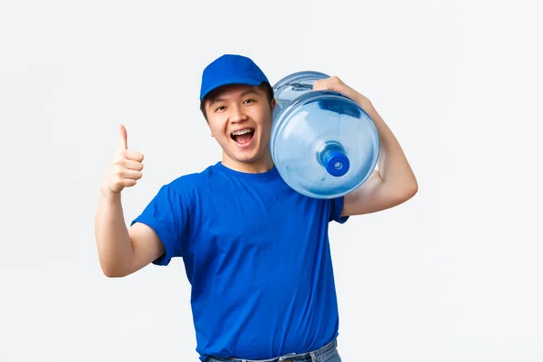 オフィスと家庭の配達と従業員の概念。青い制服の若い陽気なasain男性宅配便,肩の上にきれいな水でボトルを運ぶ,クライアントに注文をもたらし、親指を表示-アップ — ストック写真