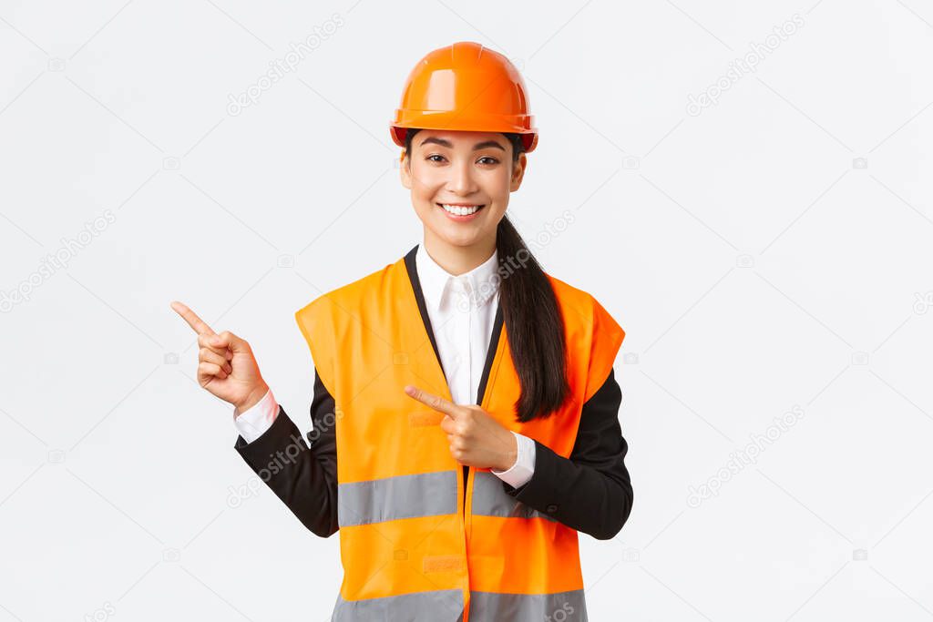 Building, construction and industrial concept. Smiling asian female architect in safety helmet, reflective clothing pointing finger upper left corner, showing project on workplace, white background