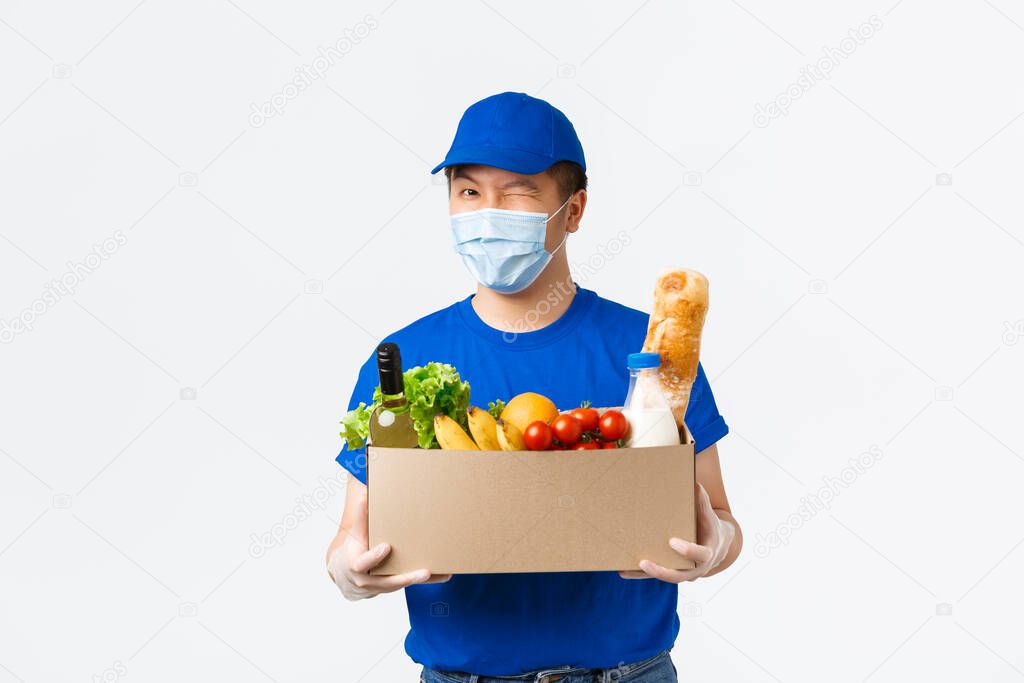 Online shopping, food delivery and covid-19 pandemic concept. Cheerful asian male courier provide fast shipping, holding box with groceries, bring order to client, wear medical mask and gloves