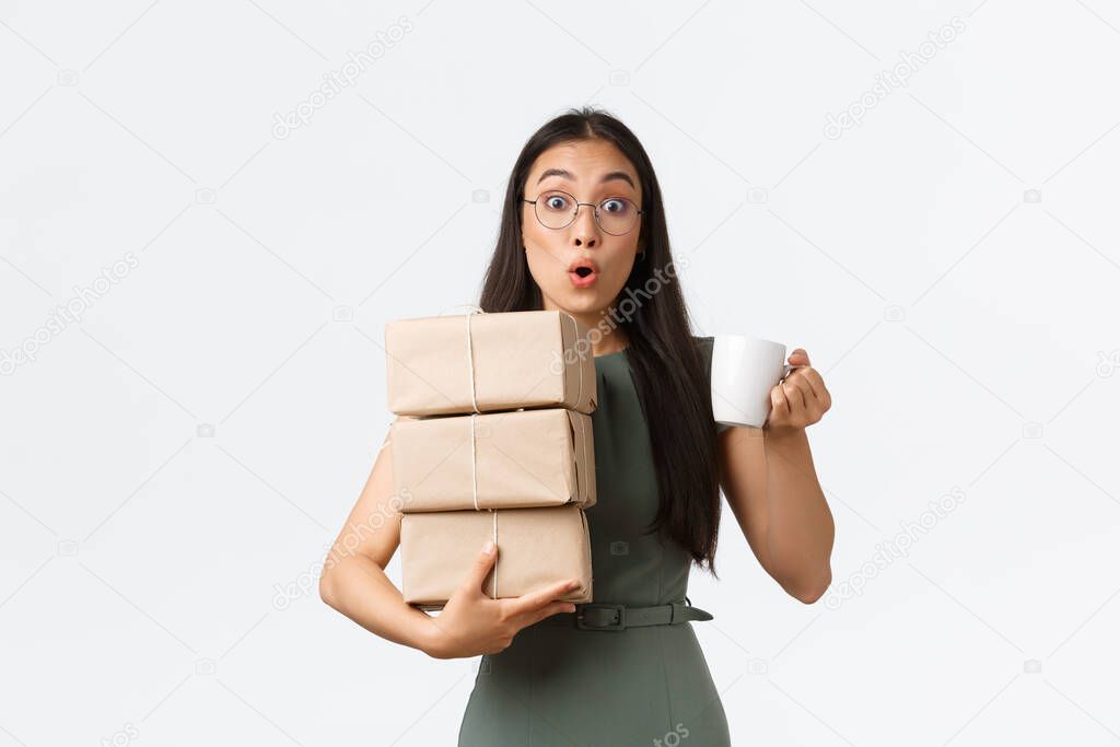 Young female asian assistant, office lady carry delivery to boss office and bring coffee mug, holding piles of boxes and cup with drink, standing white background excited
