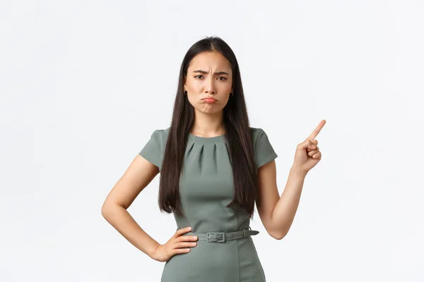 Small business owners, women entrepreneurs concept. Skeptical and disappointed asian female in dress complaining, pointing finger upper right corner at grimacing, pouting upset, white background — Stock Photo, Image