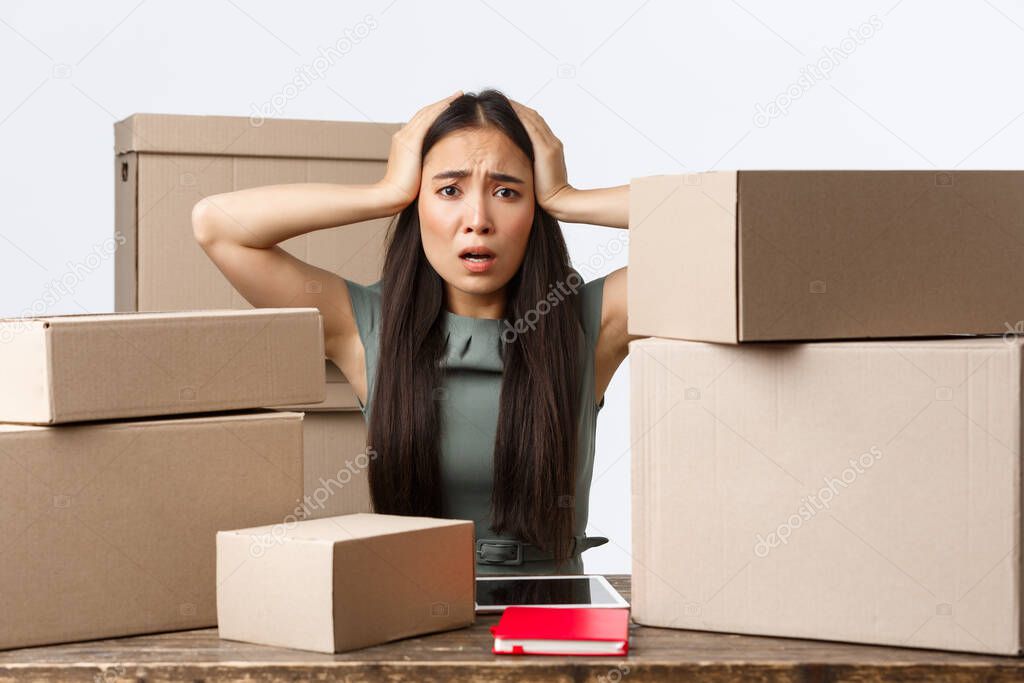 Small business owners and e-commerce concept. Worried asian businesswoman panicking, cant process all orders from her online shop, grab head anxious as sitting with lots of boxes ready for shipping