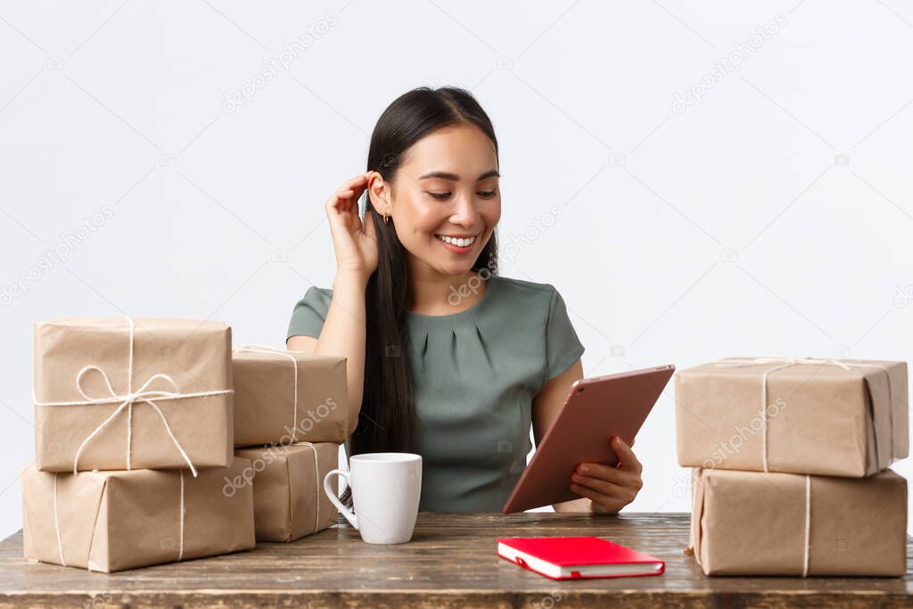 Small business owners, startup and e-commerce concept. Successful asian businesswoman with online shop, answering clients with digital tablet while drinking coffee and packign orders for shipping