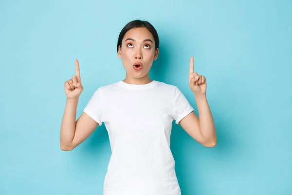 Sorprendido e impresionado chica asiática en camiseta blanca mirando y señalando los dedos hacia arriba en algo interesante y sorprendente. Hermosa chica coreana de 20 años encontró impresionante oferta promocional, sobre fondo azul — Foto de Stock