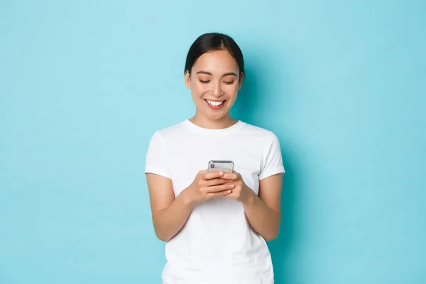Livsstil, teknik och e-handel koncept. Porträtt av söt asiatisk flicka i vit casual t-shirt ler mot mobiltelefon skärmen. Koreanska kvinna med hjälp av smartphone ansökan över blå bakgrund — Stockfoto