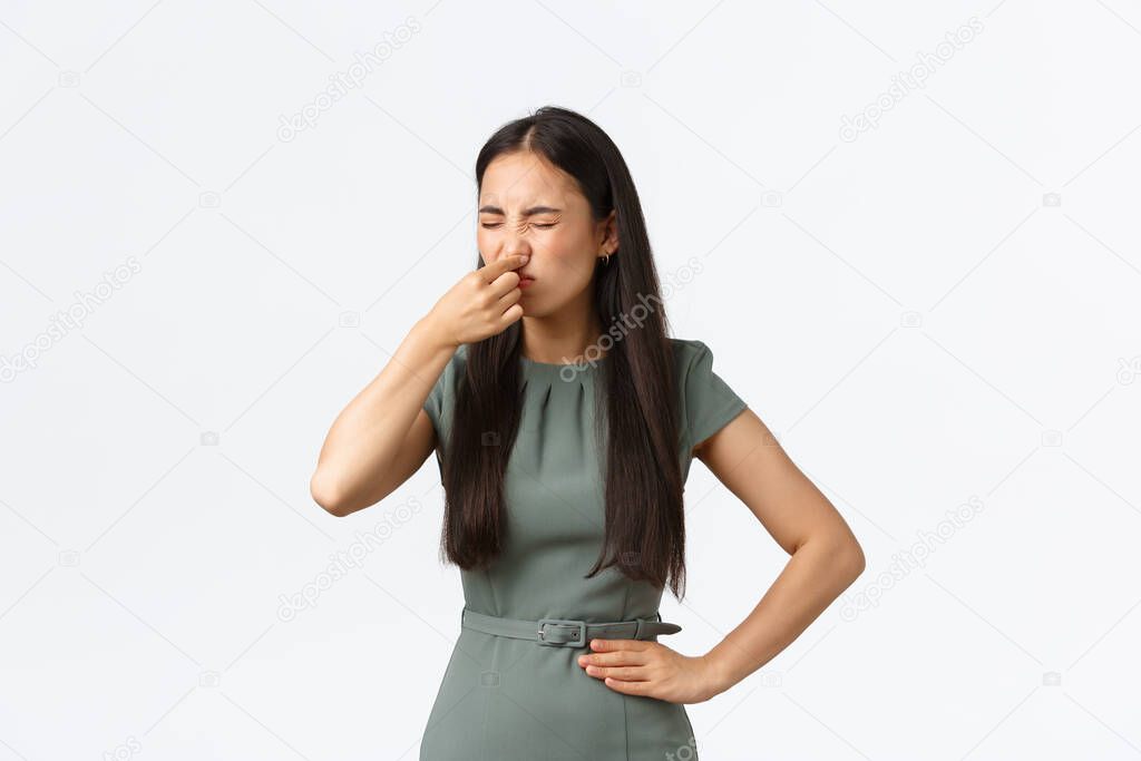Small business owners, women entrepreneurs concept. Disgusted asian female in elegant dress shut her nose with fingers and grimacing from nasty reek, bad smell, standing white background