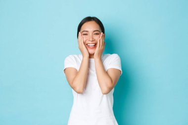 Skincare, beauty and lifestyle concept. Cheerful smiling asian girl rejoicing, looking happy, touching perfectly clean skin and rejoicing, got rid of acne, standing blue background clipart