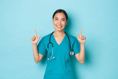 Covid-19, sosyal uzaklık ve koronavirüs salgını konsepti. Gülümseyen güzel Asyalı doktor, önlüklü hemşire mutlu bir ifadeyle parmaklarını kaldırıyor.