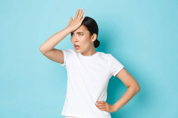 Bothered and annoyed asian girl in white t-shirt punch forehead, facepalm and roll eyes displeased, forgot something, hear dumb question, standing tensed and stressed-out blue background — Foto Stock