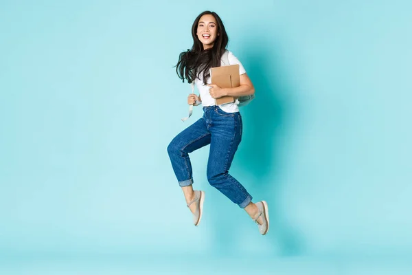 Fashion, back to school and lifestyle concept. Carefree happy asian female college student, girl jumping from happiness with backpack and notebooks over light blue background