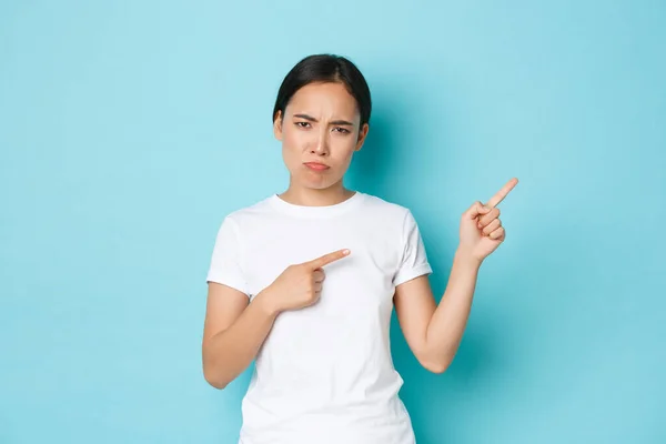 Besviken ledsen tjurande asiatisk flicka i vit t-shirt rynkar pannan och ser störd, peka finger övre högra hörnet, klaga något orättvist, svartsjuk på något, blå bakgrund — Stockfoto