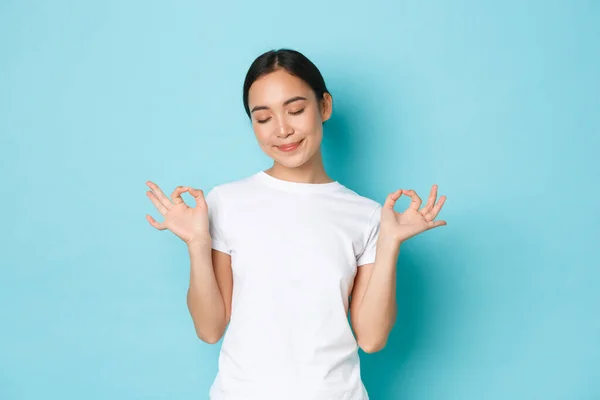 Tålamod är nyckeln. Lugn och avslappnad leende asiatisk flicka med slutna ögon och lotus utgör, står över blå bakgrund, meditera eller släppa stress, ser fridfull och glad — Stockfoto