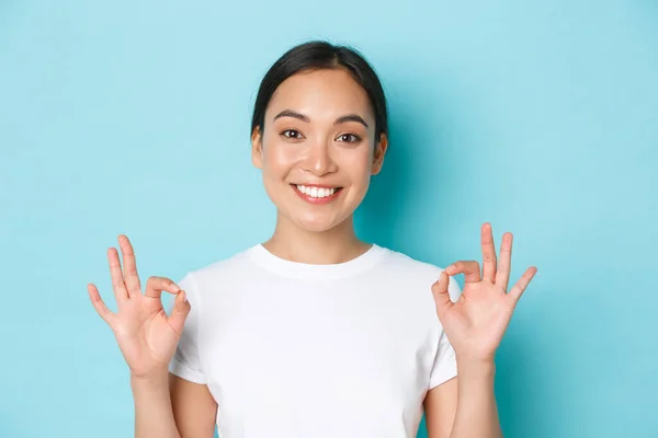 Primer plano de bastante satisfecho sonriente chica asiática en camiseta blanca, mostrando gesto de recomendación, signo de bien y asentir con la cabeza en la aprobación, elección de cumplido, de acuerdo con usted, de pie fondo azul claro —  Fotos de Stock