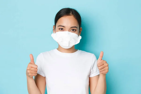 Covid-19, distanciamento social e conceito de pandemia de coronavírus. Close-up de confiante e assertiva menina asiática em t-shirt branca e respirador médico mostrando polegares para cima, fundo azul claro — Fotografia de Stock