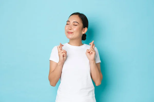 Cintura-up de sonhador esperançoso, bela menina asiática cruz dedos boa sorte e fechar os olhos ao fazer desejo, sorrindo otimista, orando para o sonho se tornar realidade, de pé fundo azul — Fotografia de Stock