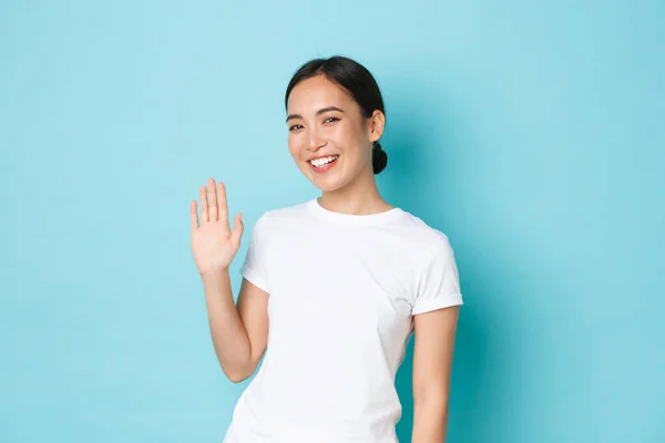 Amigável-olhando alegre menina asiática dizendo Olá, cumprimentando novas pessoas na companhia, Sorrindo coreano feminino acenando mão para dizer oi, bem-vindo alguém, de pé fundo azul otimista — Fotografia de Stock