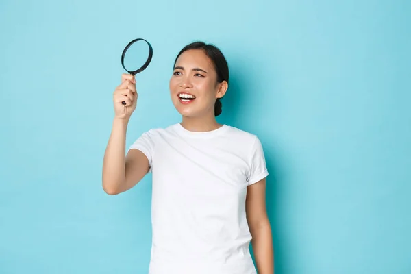 Porträtt av rolig och söt asiatisk kvinna i vit t-shirt, söker efter något, tittar genom förstoringsglas för att läsa små bokstäver, squinting förbryllad, stående ljusblå bakgrund — Stockfoto