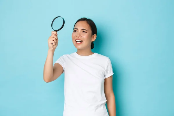 Retrato de engraçado e bonito asiático fêmea em branco t-shirt, à procura de algo, olhando através de lupa para ler letras pequenas, squinting perplexo, de pé luz azul fundo — Fotografia de Stock