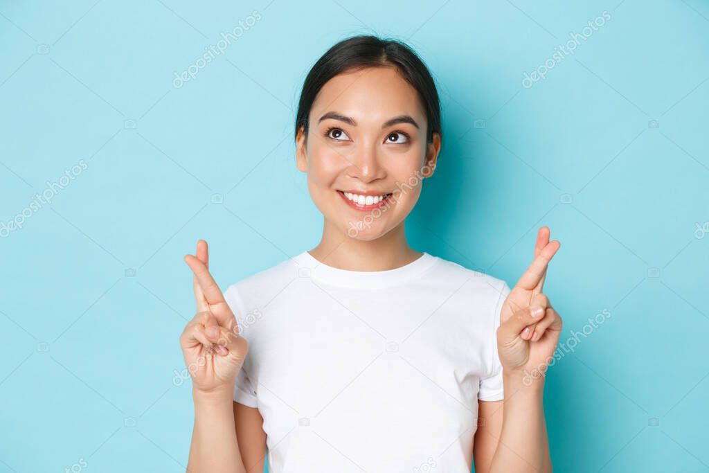 Close-up of hopeful dreamy asian girl making wish, looking upper left corner thoughtful, standing light blue background with fingers crossed for good luck, praying or anticipating positive news