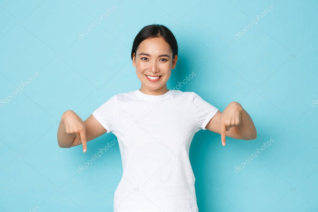 Portrait of confident beautiful asian girl in white t-shirt, pointing fingers down, showing way, demonstrating banner with advertisement over blue background. Korean woman promote product