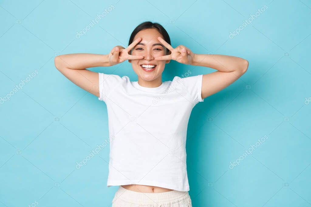 Lifestyle, beauty and women concept. Cheerful beautiful asian girl in white t-shirt looking upbeat and kawaii, showing peace gesture over eye and smiling broadly, standing blue background