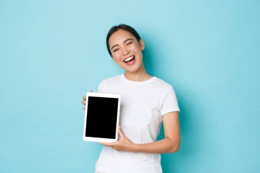 Gururlu ve mutlu gülümseyen Asyalı kız portresi, sevinçle sırıtan, dijital tablet ekranını gösterirken mutlu görünen, iş projesini veya reklamını gösteren, açık mavi arka plan