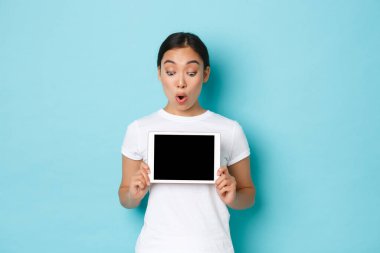 Heyecanlı ve şaşırmış beyaz tişörtlü güzel Asyalı kadın dijital tablet ekrana bakıyor, şaşırmış, heyecanlanmış bir şekilde, grafik gösteriyor veya online alışveriş sitesini gösteriyor, açık mavi arka plan