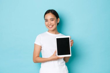 Gülümseyen çekici Asyalı kız portresi dijital tablet ekranda proje veya reklam sunuyor, yeni alışveriş sitesi tanıtıyor, açık mavi arka planda neşeli duruyor