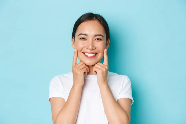 Güzel Asyalı kız mutlu görünüyor, gülümsüyor beyaz dişler, yanaklara dokunuyor, cilt bakım ürünleri öneriyor, yüz bakımı için kozmetik ürünleri, açık mavi arka plan — Stok fotoğraf