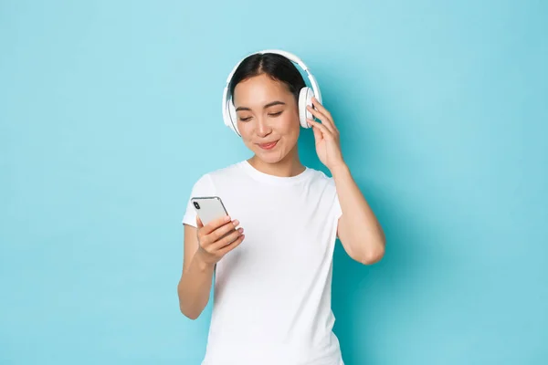 Lifestyle, people and leisure concept. Smiling attractive asian woman picking song in playlist, looking at smartphone screen while deciding what listen in headphones, blue background — Stock Photo, Image