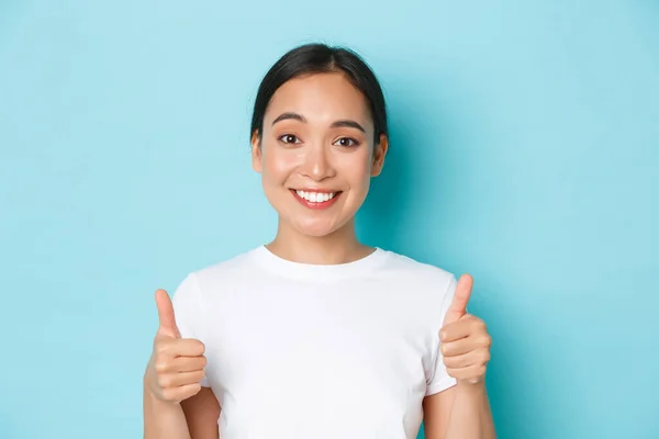 Primer plano de satisfecha y alegre sonriente chica asiática en camiseta blanca, mostrando los pulgares hacia arriba de apoyo y buscando complacido en la cámara, recomendar el producto, felicitar a la buena opción —  Fotos de Stock