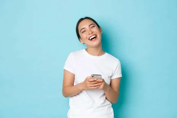Livsstil, teknik och e-handel koncept. Porträtt av bekymmerslös glad asiatisk flicka skrattar och ler efter att ha läst något roligt på nätet, hålla smartphone och ha kul, blå bakgrund — Stockfoto