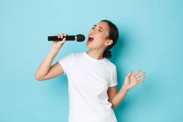 Styl życia, ludzie i pojęcie czasu wolnego. Namiętna i beztroska ładna azjatycka dziewczyna śpiewająca piosenkę w mikrofonie, zginająca się podczas występu, jak karaoke, stojące jasnoniebieskie tło wykonujące — Zdjęcie stockowe