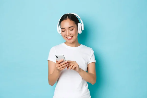 Lifestyle, people and leisure concept. Smiling attractive asian female using wireless headphones and mobile phone, watching video at crowded place, listening music, light blue background — Stock Photo, Image