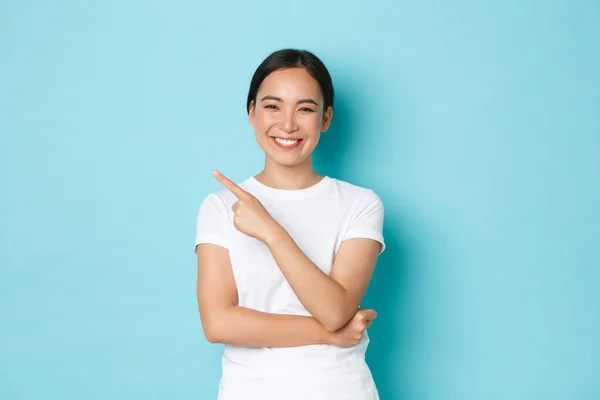 Fröhlich selbstbewusstes asiatisches Mädchen im weißen T-Shirt, mit dem Finger in die linke obere Ecke zeigend und entzückt lächelnd, den Weg zeigend, Werbeangebot. Studentin trifft ihre Wahl und demonstriert es — Stockfoto